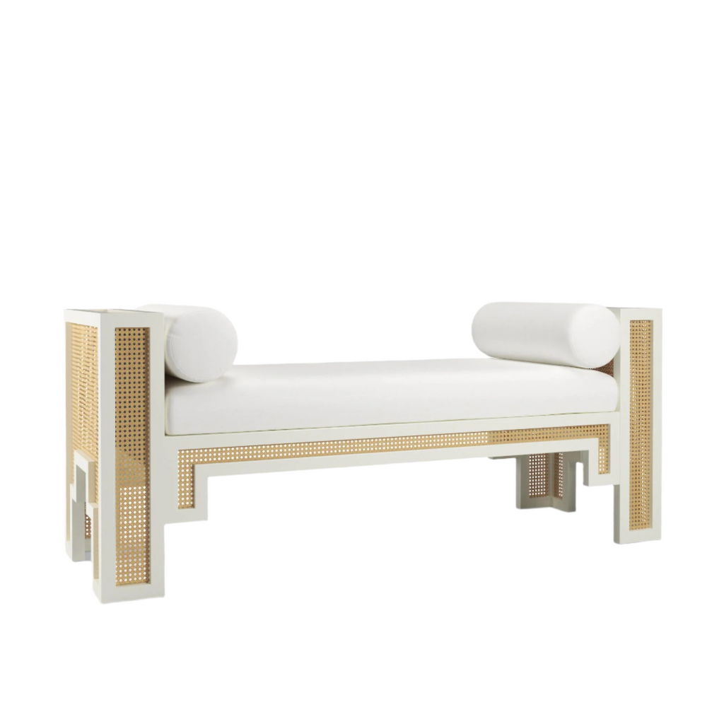 White upholstered end-of-bed bench with natural rattan cane panels set into a white solid wood frame, geometric cutout legs and two cylindrical bolster pillows in a boho-Scandinavian style.
