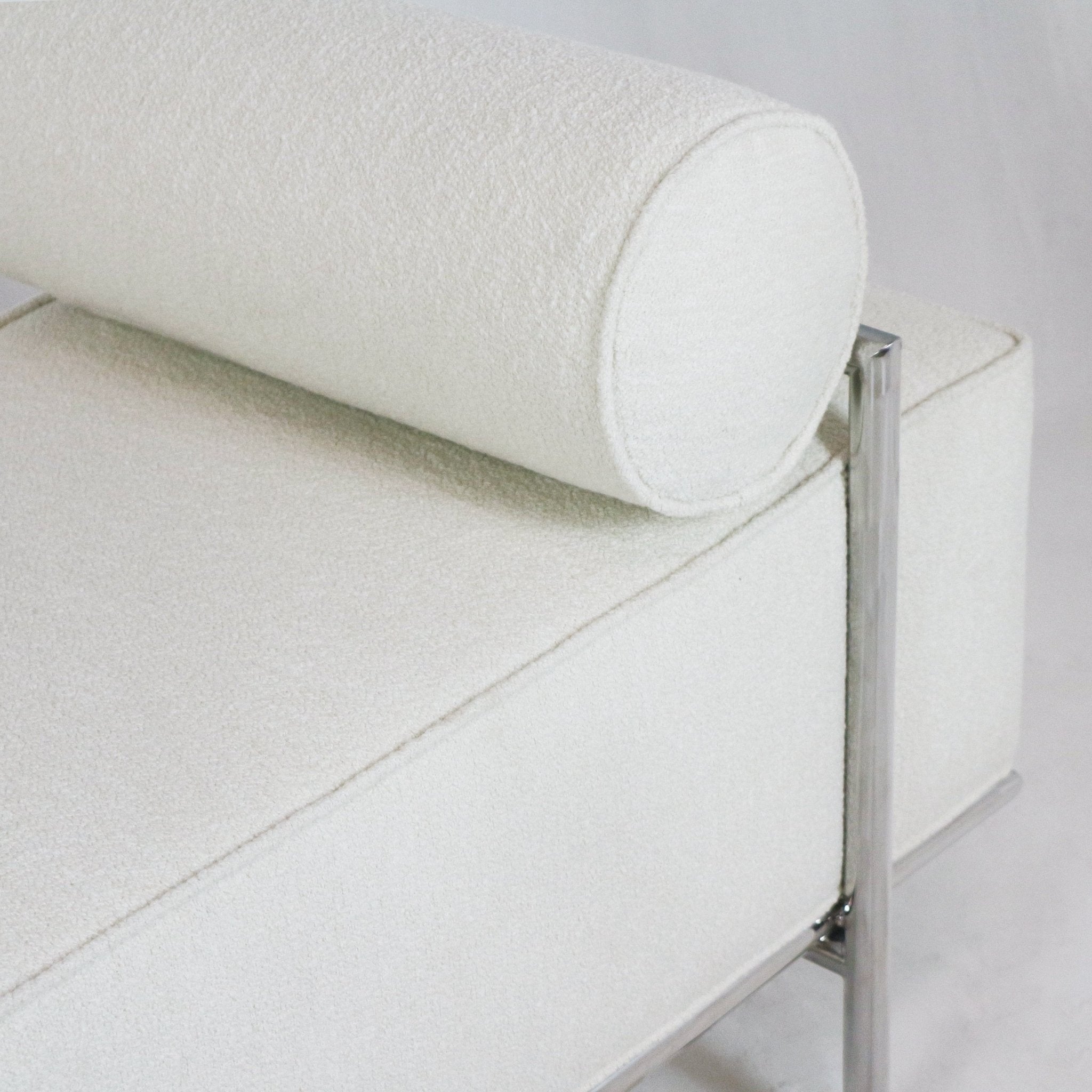Close-up of a white boucle upholstered end-of-bed bench showing a round bolster pillow, cushioned rectangular seat, and polished chrome metal leg and frame.
