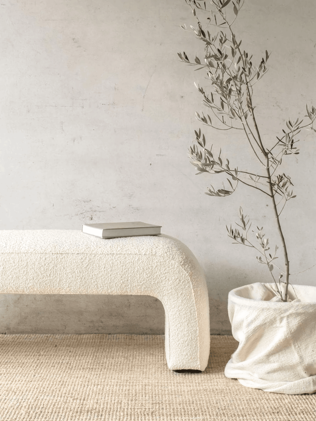 Cream boucle upholstered arched end-of-bed bench with a closed book on top, placed on a natural jute rug against a textured plaster wall, beside a potted olive branch in a soft linen-like fabric basket.