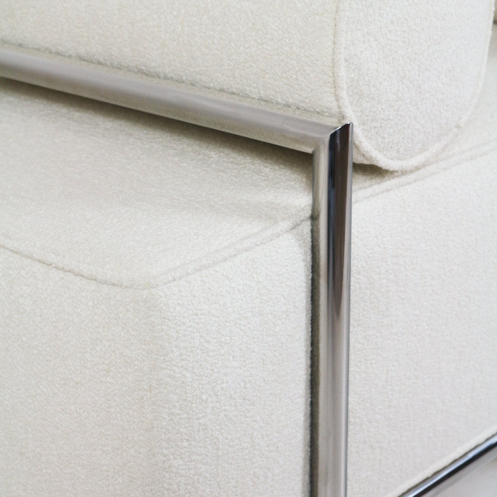 Close-up of a cream boucle-upholstered end-of-bed bench showing a piped seat edge and a polished stainless-steel tubular frame and leg in a modern, minimalist style.