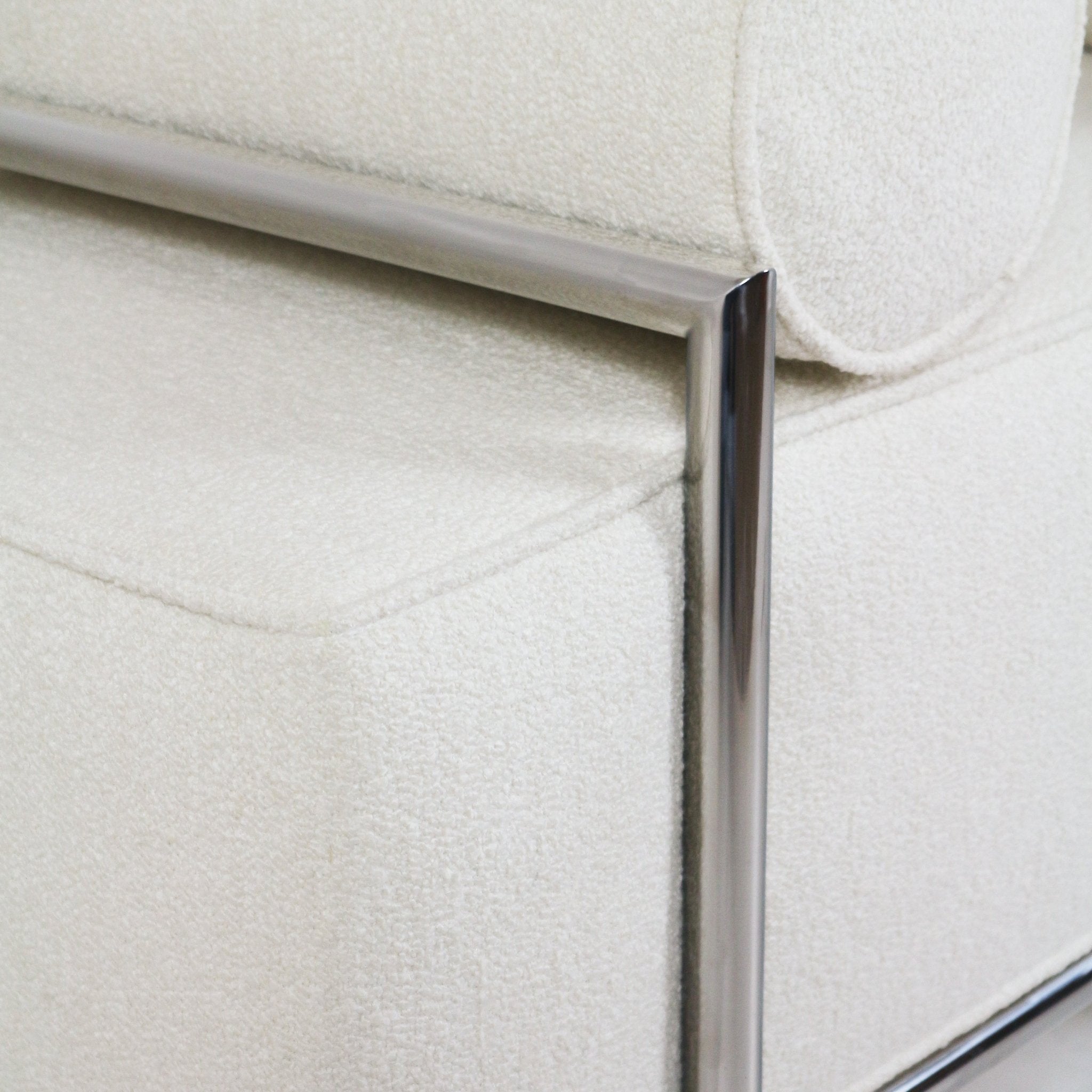 Close-up of a cream boucle-upholstered end-of-bed bench showing a piped seat edge and a polished stainless-steel tubular frame and leg in a modern, minimalist style.