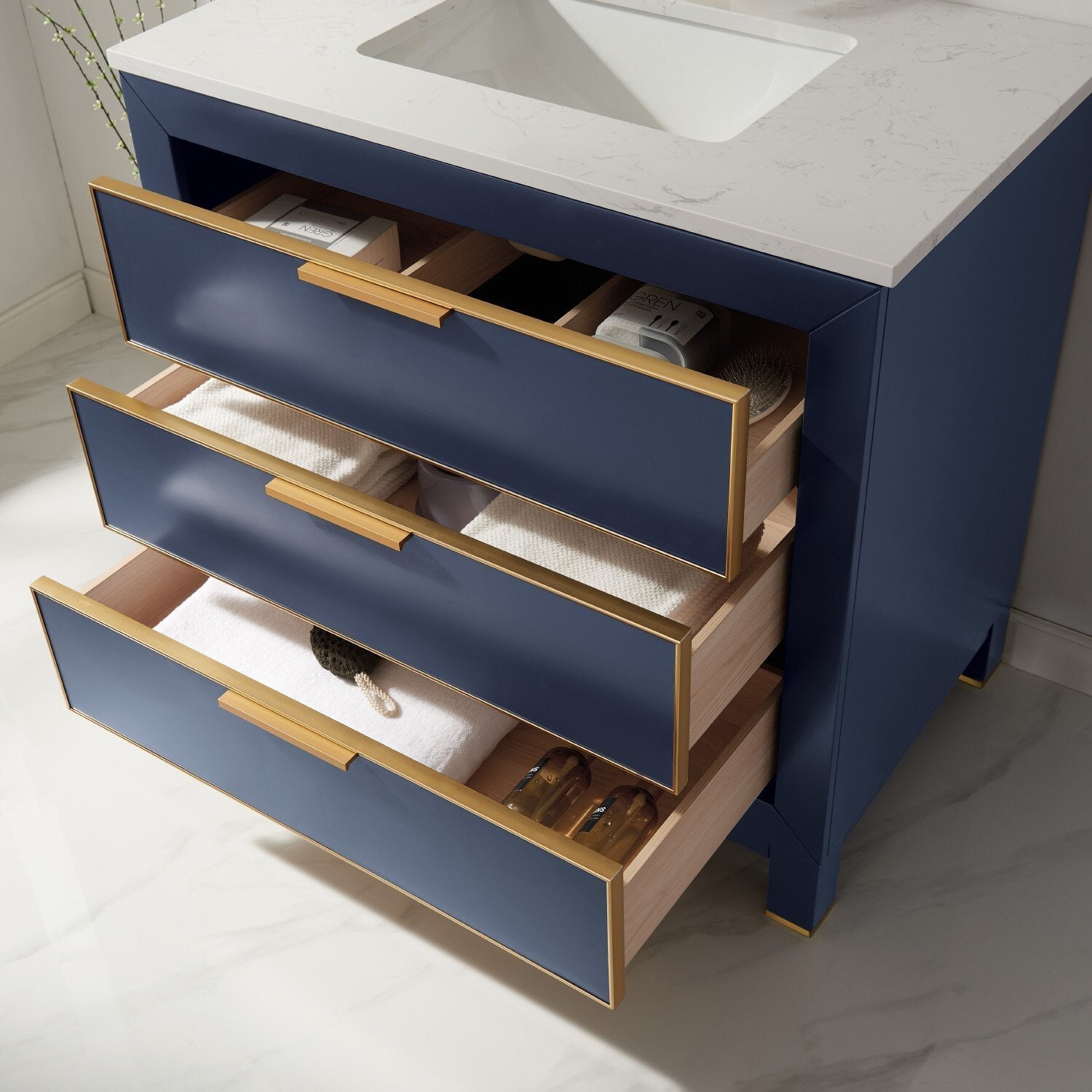 Freestanding 36-inch modern bathroom vanity with a white stone top and integrated ceramic sink, navy blue drawer fronts with gold trim and handles, three open drawers showing solid wood interiors and folded towels.