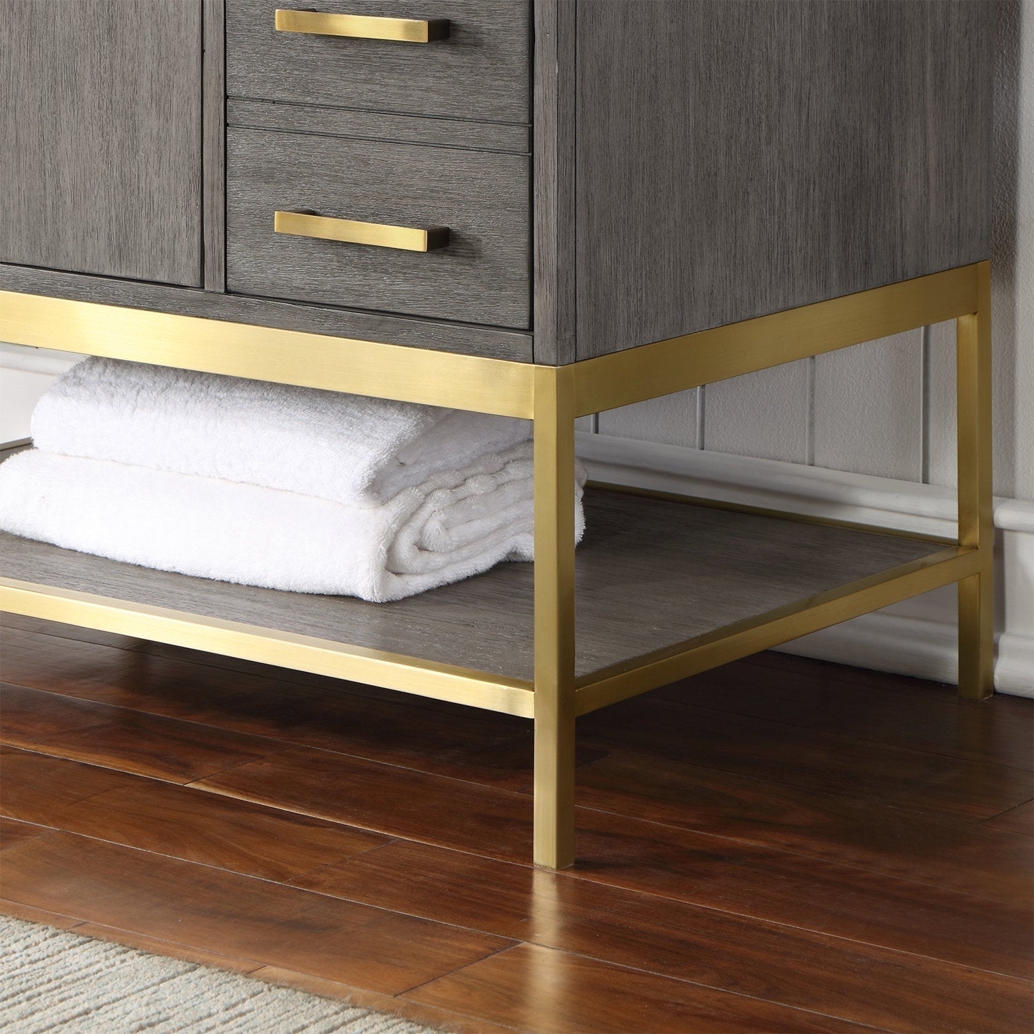 Close-up of a stone-colored solid wood bathroom vanity corner with a brass-finished metal base and matching drawer pulls; lower open shelf holds folded white towels above a hardwood floor.