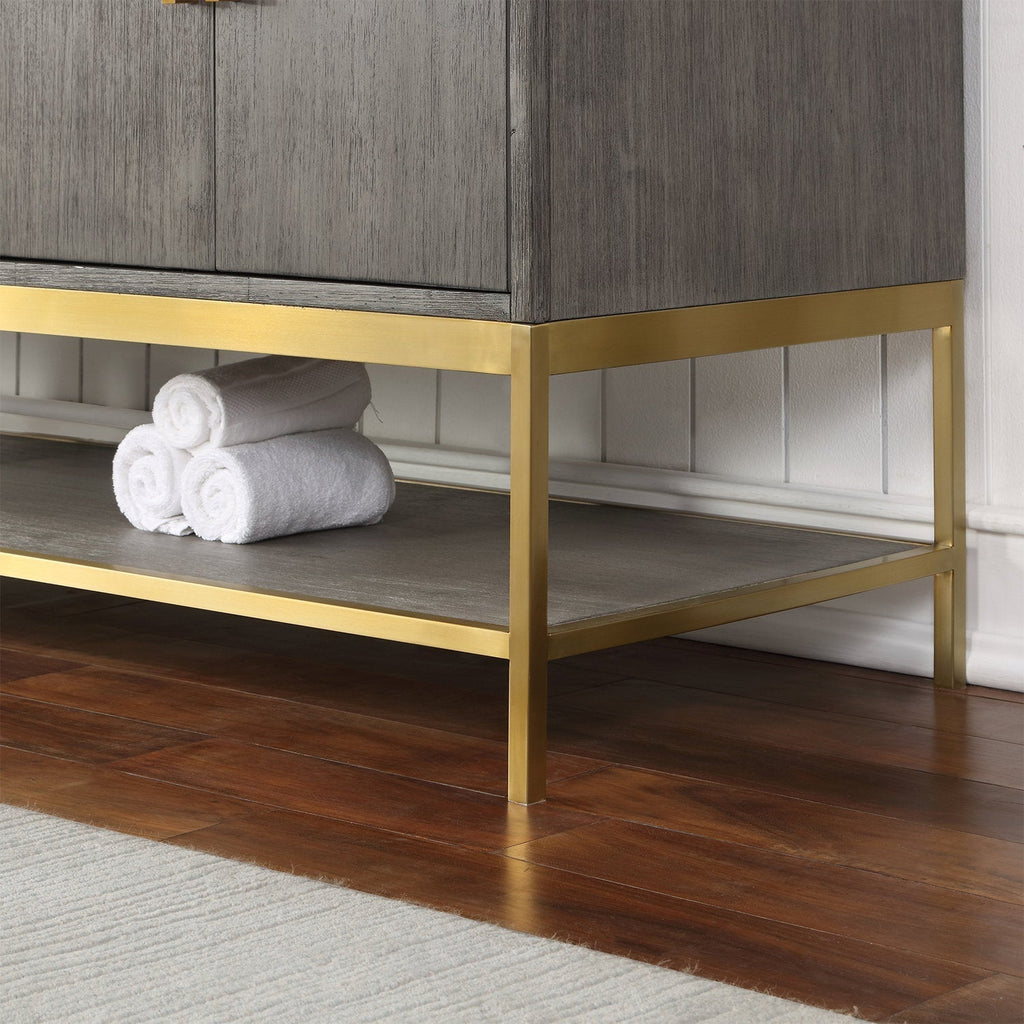 Close-up of the lower corner of a modern solid wood bathroom vanity in a stone finish with a gold-finished metal base and open shelf holding rolled white towels on a hardwood floor.