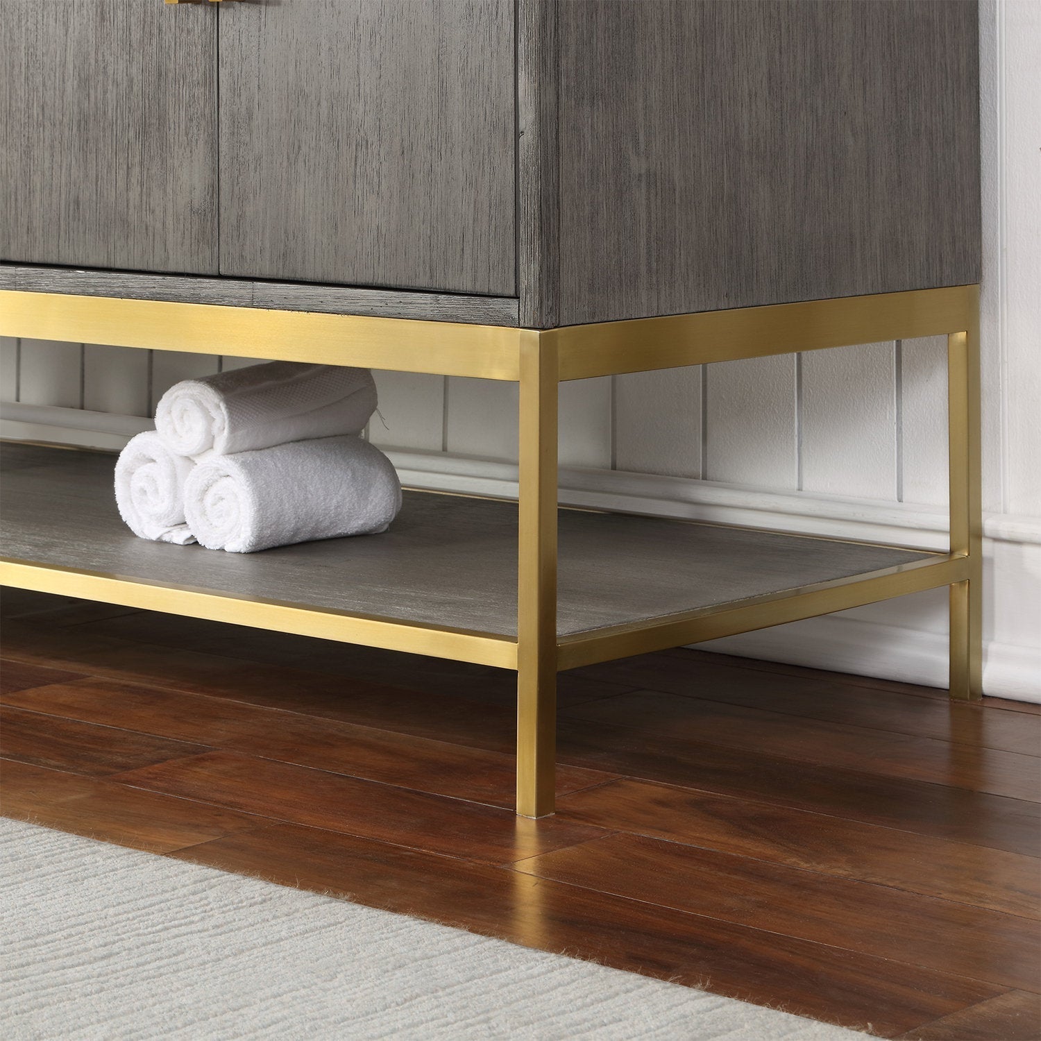 Close-up of the lower corner of a modern solid wood bathroom vanity in a stone finish with a gold-finished metal base and open shelf holding rolled white towels on a hardwood floor.