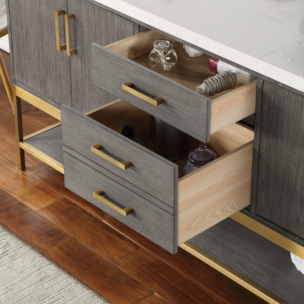 Close-up of a modern solid wood bathroom vanity in stone finish with a white stone countertop, two open wood-lined drawers, gold handles and a gold metal base holding bathroom accessories.