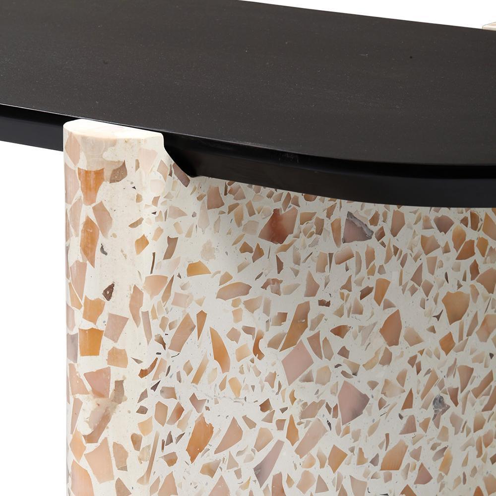 Close-up of a modern coffee table showing a black wooden tabletop edge resting on a rounded terrazzo stone base with a cream background and warm beige, pink and amber chips.