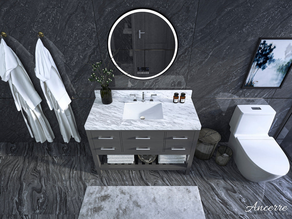 Contemporary 48-inch solid wood bathroom vanity with a white marble top and rectangular undermount sink, gray base with six drawers and open towel shelf, round illuminated mirror, dark stone walls and a white toilet nearby.