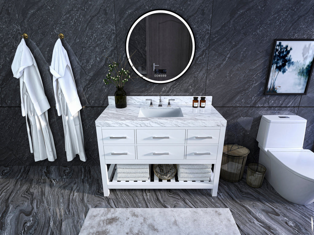 Freestanding white solid-wood single-sink vanity with a marble top and rectangular undermount sink, brushed metal drawer pulls and open towel shelf; round illuminated mirror on a dark stone wall with robes and a toilet nearby.