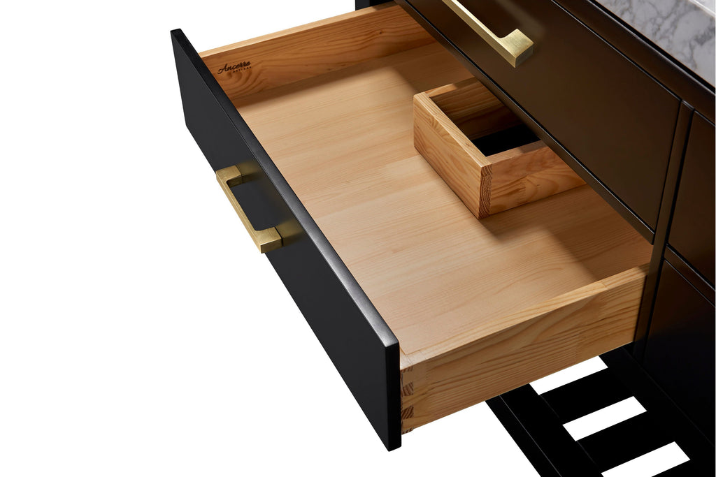 Open black bathroom vanity drawer revealing light solid wood interior with dovetail joints and a small wooden organizer, brushed gold metal handle on the matte black face and a glimpse of stone countertop and slatted shelf.