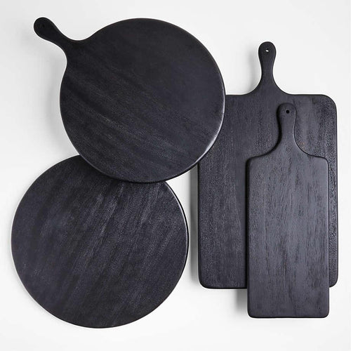 Overhead view of four black ash solid wood cutting and serving boards — two round paddle boards and two rectangular paddles with handles — arranged on a white background in a modern, minimalist style.
