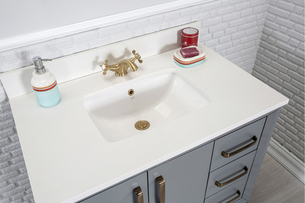 Modern gray solid-wood single-sink vanity with a white stone countertop and integrated ceramic sink, brass faucet and hardware, soap dispenser and dish on a white brick backsplash.