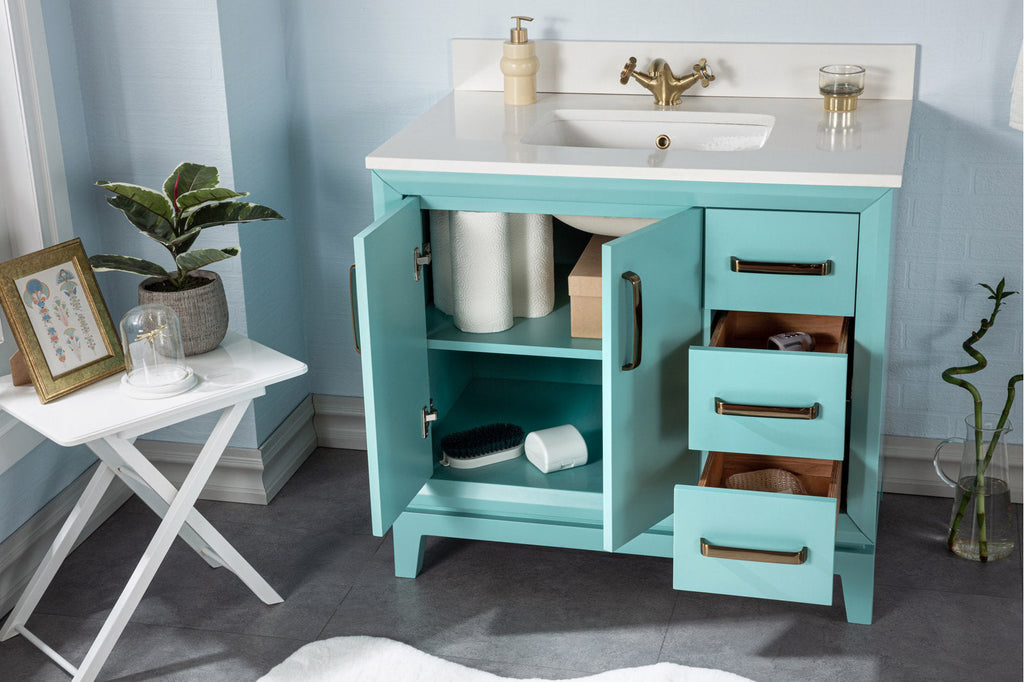 Modern turquoise solid-wood bathroom vanity with a white stone top and brass-tone handles; freestanding single-sink unit shows open cabinet doors and three drawers revealing shelving with toilet paper and grooming accessories.