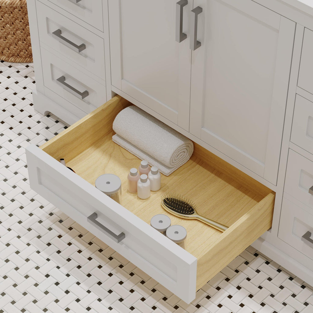 Open modern stone-colored bathroom vanity drawer with solid wood interior, containing a rolled white towel, small toiletry bottles and jars, and a gold-handled hairbrush; white shaker drawer front with metal pull over mosaic tile floor.