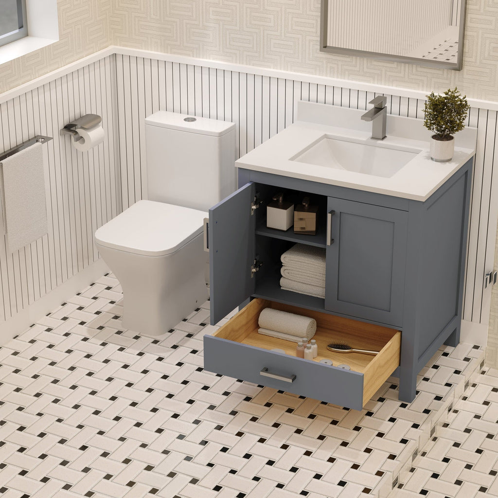 Freestanding modern solid wood vanity in blue-gray with a white stone countertop and integrated rectangular sink; open cabinet and pulled wooden drawer reveal folded towels, toiletries and a hairbrush next to a toilet on patterned tile.