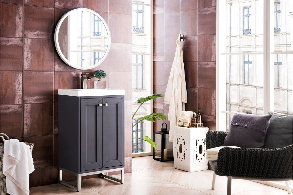 Freestanding modern solid wood bathroom vanity in a stone finish with a white stone sink top, brushed-nickel faucet and metal base, shown with a round mirror, textured brown tile wall, towel basket and decorative accessories.