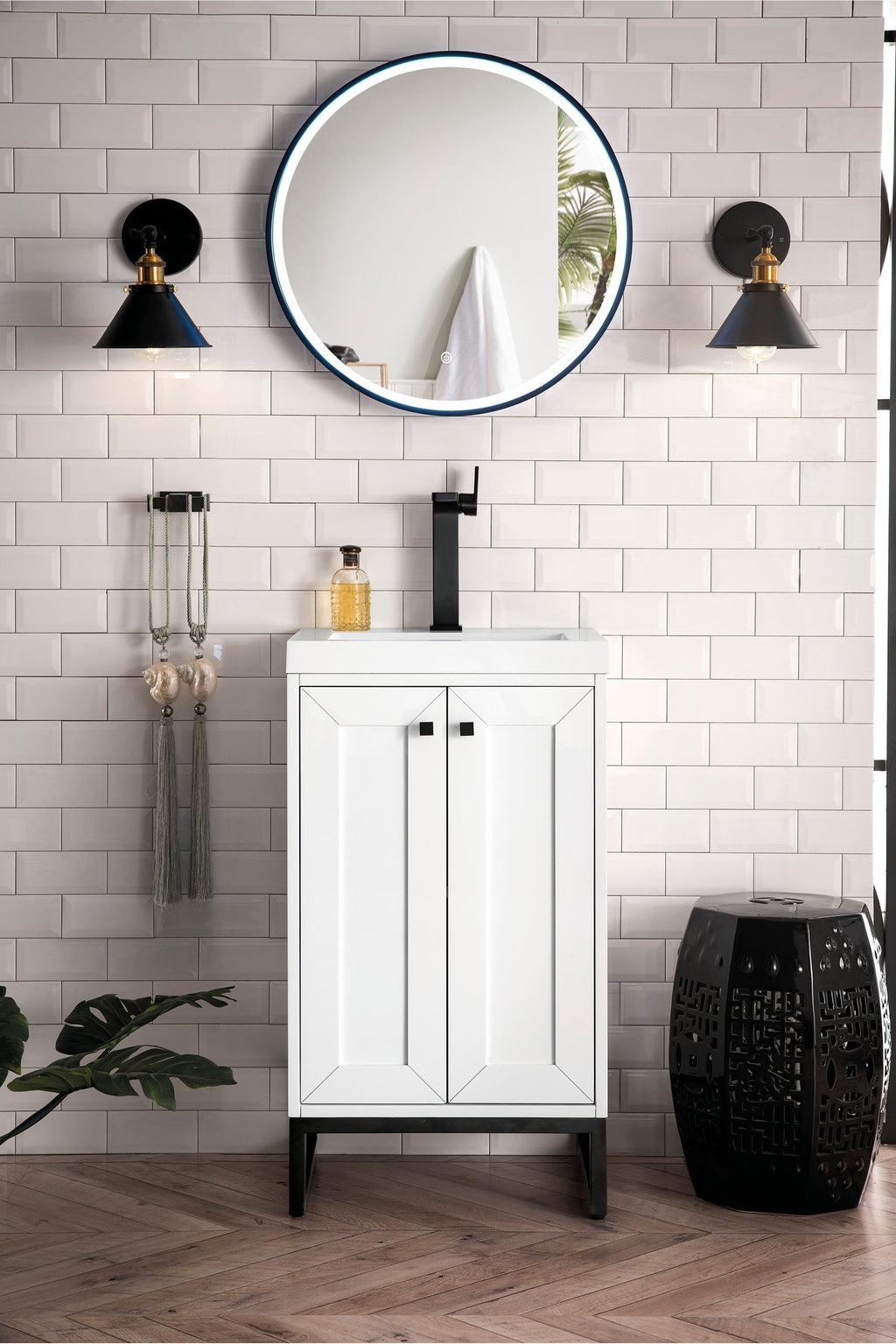 Modern white solid-wood single-sink bathroom vanity with a stone top and matte black faucet, shown with a round illuminated mirror, black wall sconces, subway tile wall and a decorative black stool.