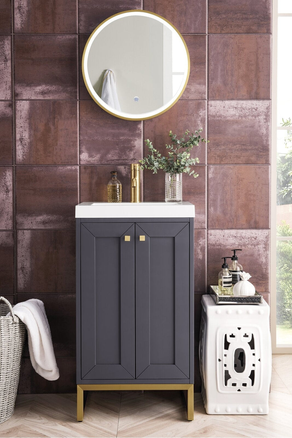 Contemporary solid-wood single-sink vanity in dark gray with a white stone top, brass faucet and gold legs, set against rust-toned textured tile with a round gold mirror and decorative vase and soap dispensers.