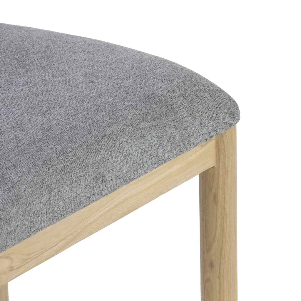 Close-up of a gray boucle upholstered seat showing textured fabric on the cushion and the natural oak apron and leg of a coastal-contemporary bar/counter stool.