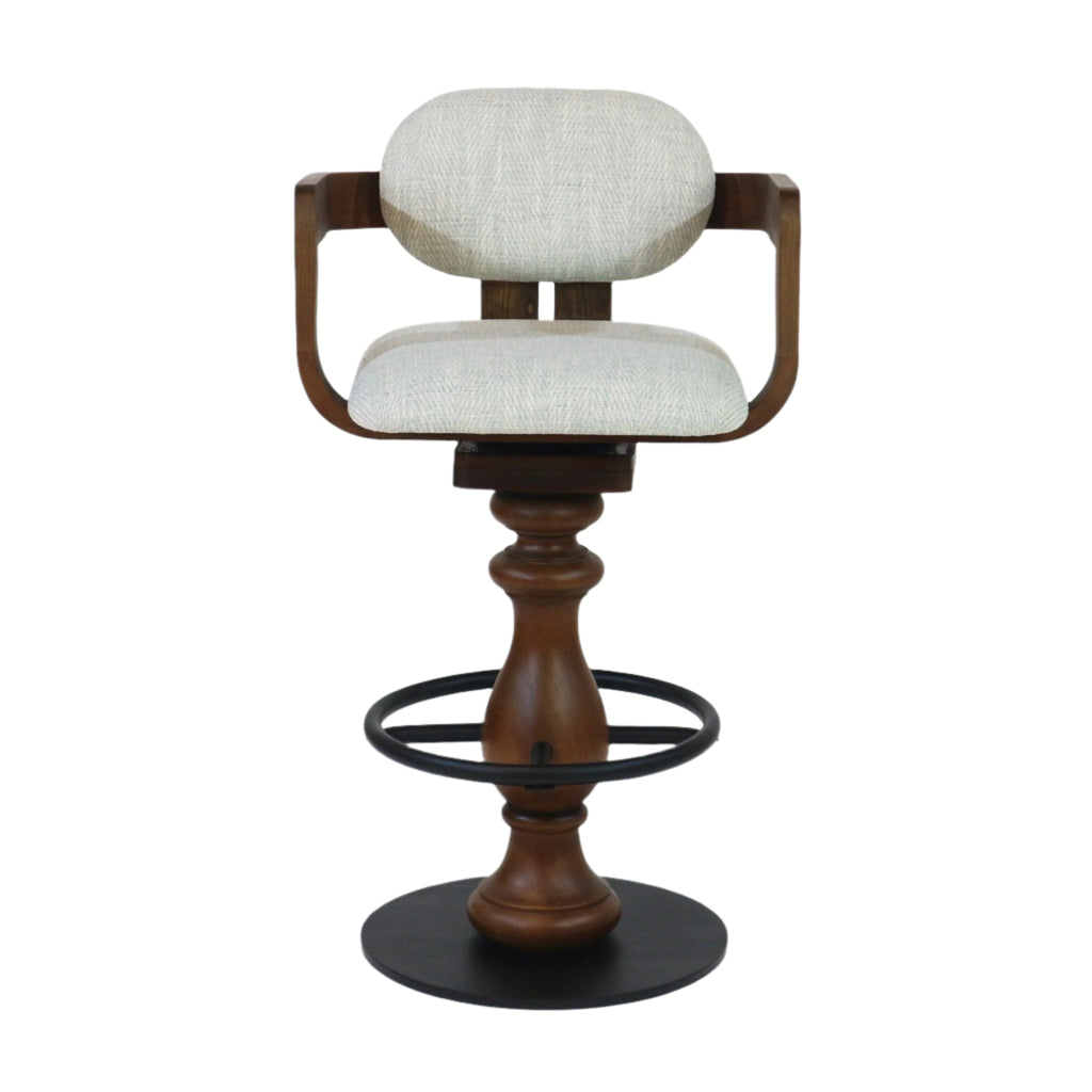 Front view of a light gray fabric-upholstered bar stool with curved wooden arms and oval cushioned back, turned natural wood pedestal, black metal circular footrest ring and round base in a counter-height swivel design.