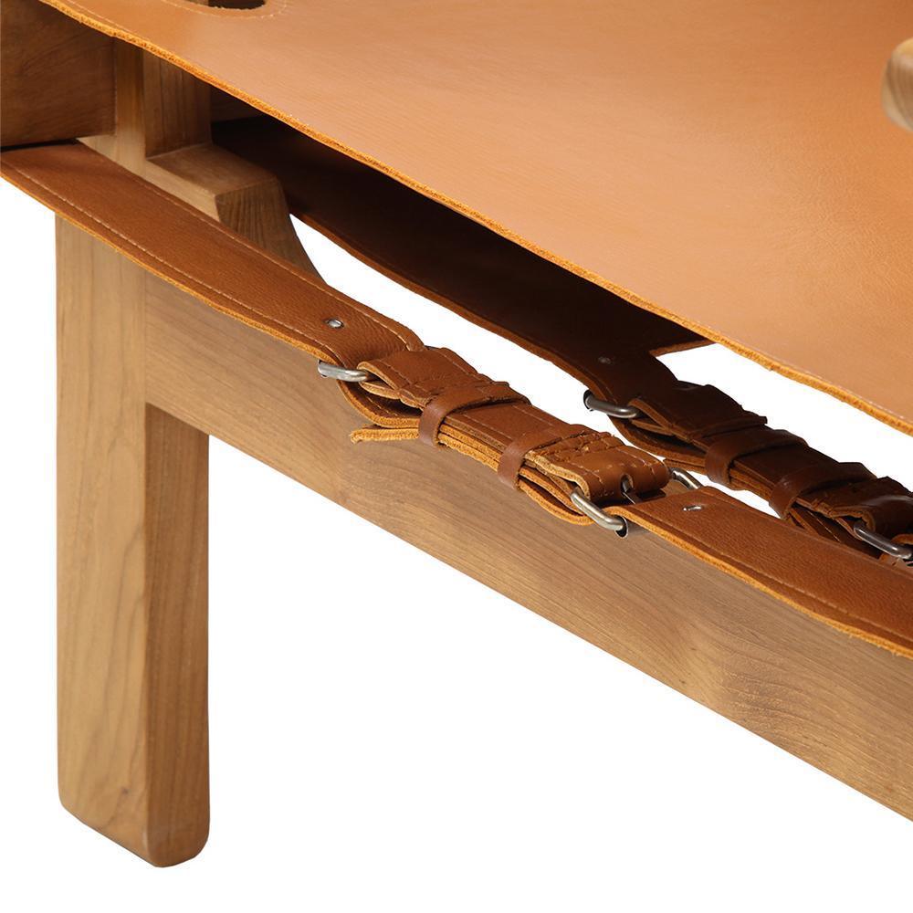 Close-up of an armchair's solid teak leg and natural tan leather seat, showing stitched leather straps and metal buckles securing the seat to the wooden frame.
