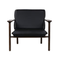 Front view of a modern mid-century lounge chair with black faux leather seat and back cushions on a warm solid wood frame with exposed armrests and round legs.