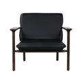 Front view of a modern mid-century lounge chair with black faux leather seat and back cushions on a warm solid wood frame with exposed armrests and round legs.