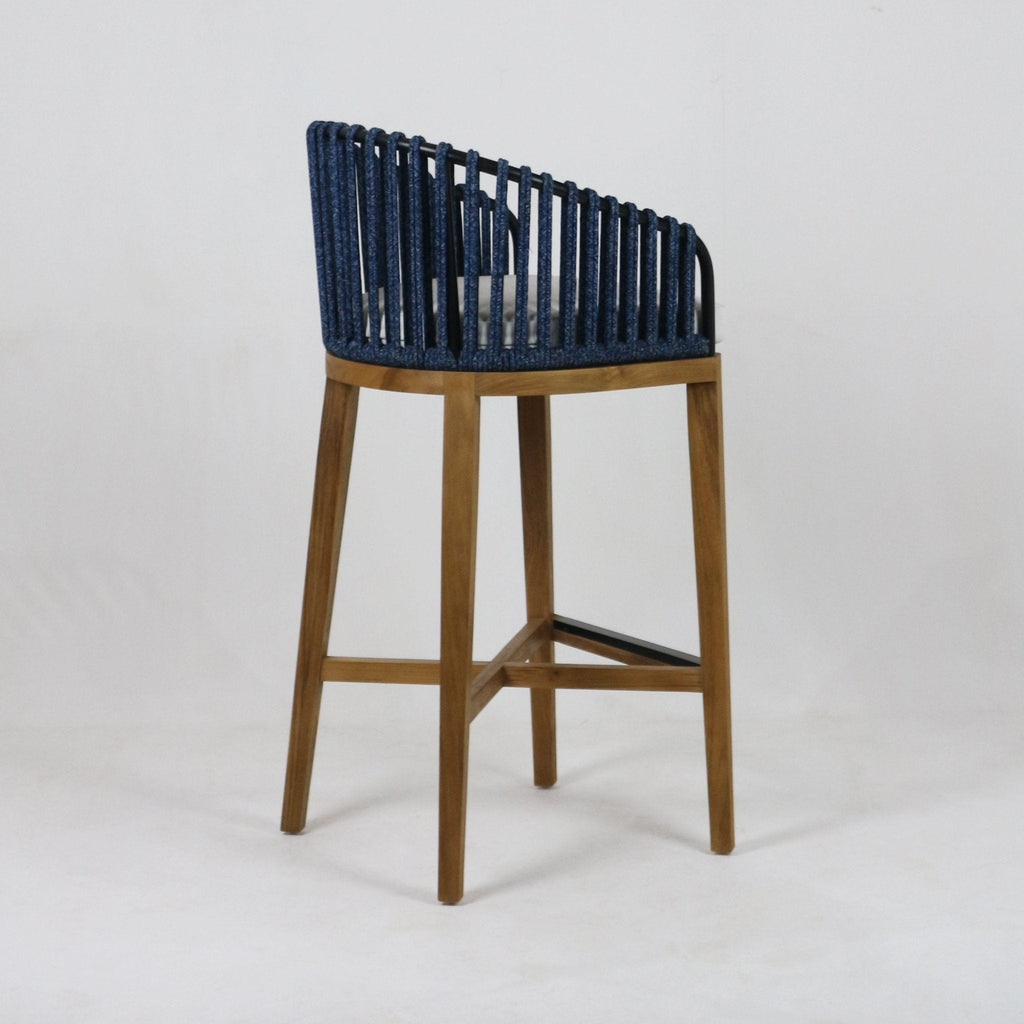 Side view of a teak-framed outdoor bar stool with a blue woven rope back, grey seat cushion, tapered natural wood legs and a black metal footrest.