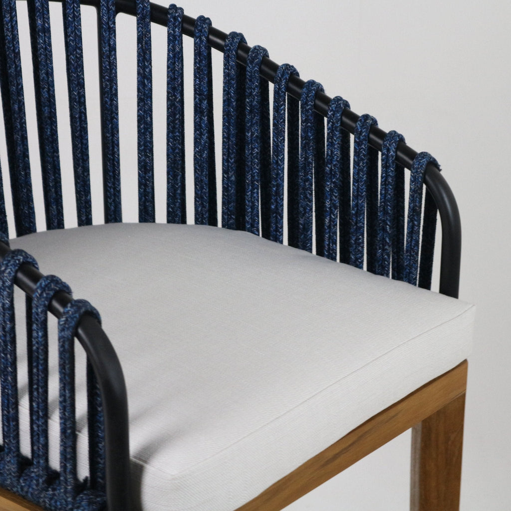 Close-up of an outdoor bar stool seat showing a light gray upholstered cushion on teak legs, a black metal frame and navy braided rope woven backrest.