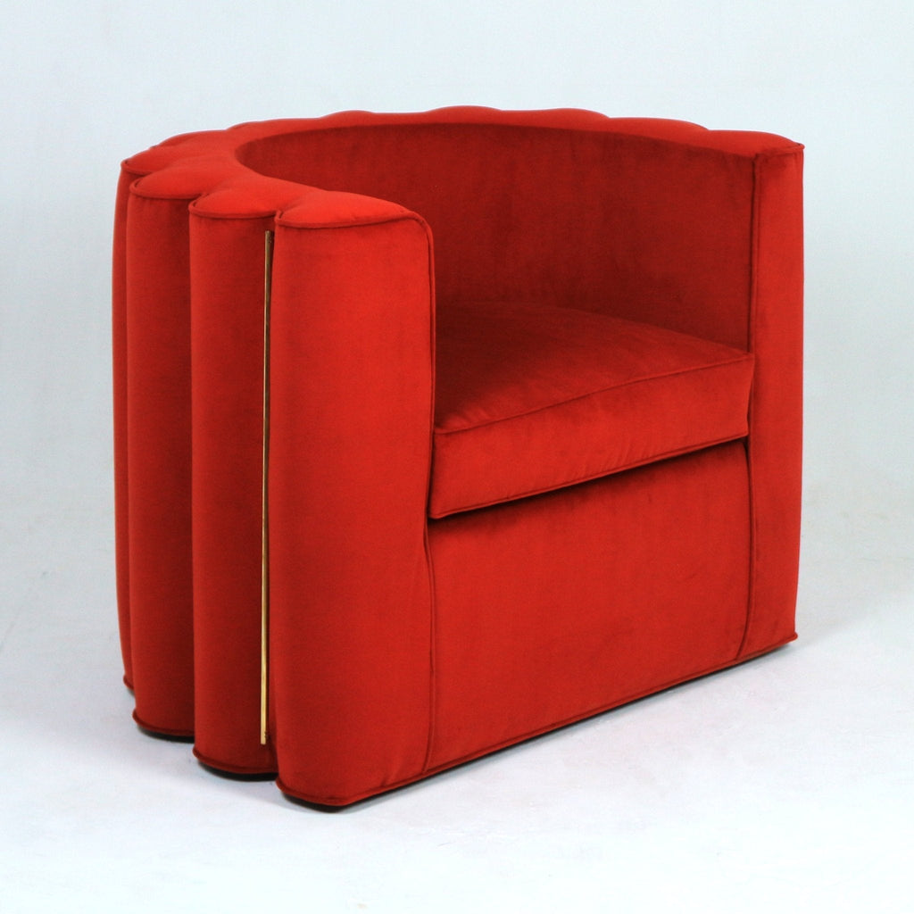 Contemporary red fabric accent chair with a rounded, scalloped full back, plush seat cushion, slim vertical gold metal trim between pleated side panels and a low dark wooden base, shown on a white background.