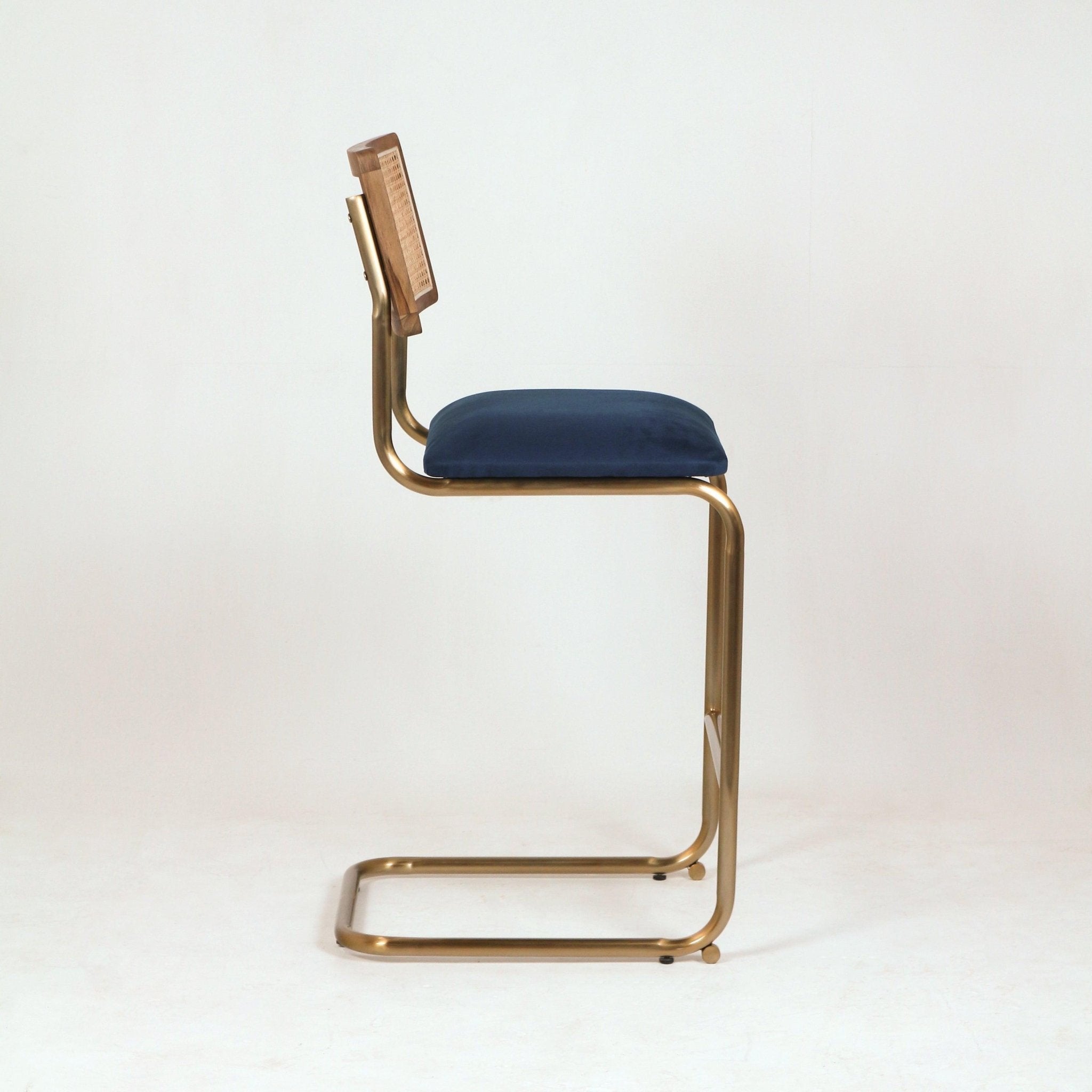Side view of a modern cantilever bar stool with a brass metal frame, navy velvet seat cushion and wood-framed woven cane back against a white background.