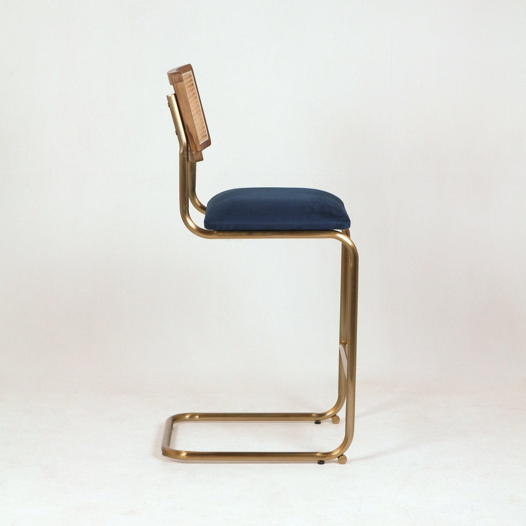 Side view of a modern cantilever bar stool with a brass metal frame, navy velvet seat cushion and wood-framed woven cane back against a white background.