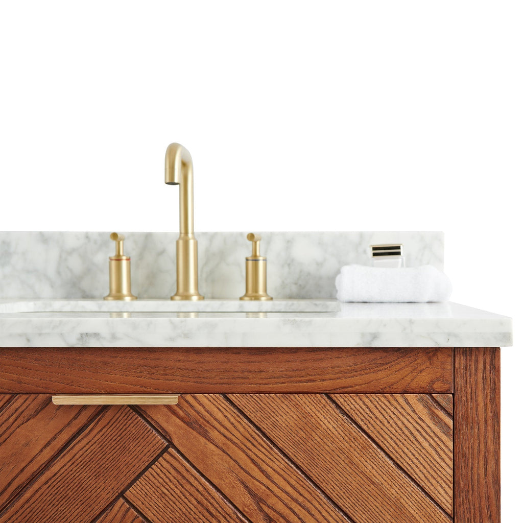 Close-up of a stone marble countertop and backsplash with brushed brass faucet and matching disc handles, a folded white towel, and a warm solid wood vanity front with diagonal chevron grain and slim brass pull.