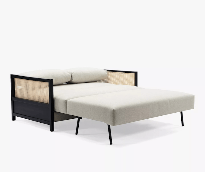 Modern beige upholstered sofa bed in bed position with two loose back cushions, a black wooden frame, woven cane side panels and slim black metal legs.