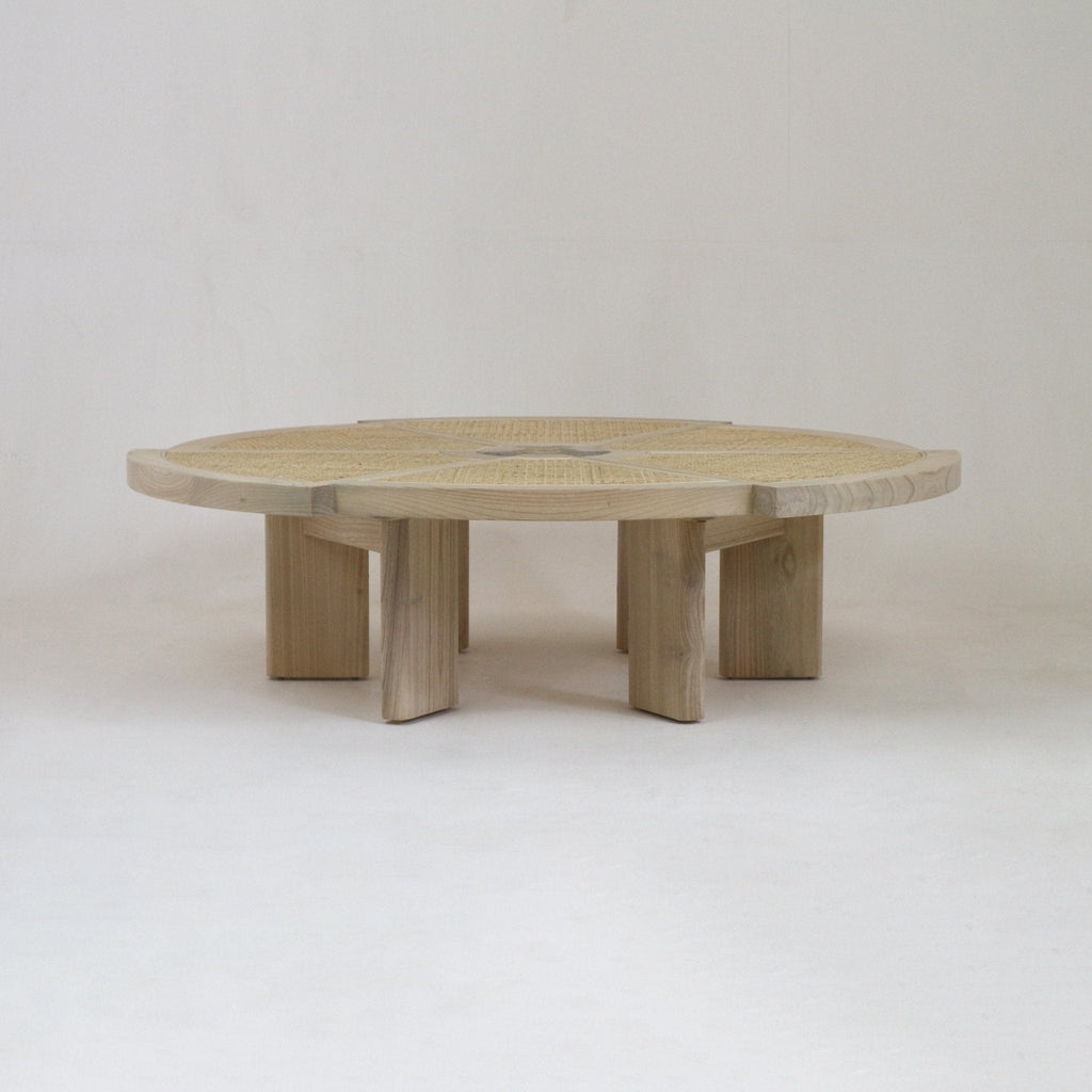 Low round coffee table in natural blonde oak with a woven cane inset top and chunky segmented pedestal legs, photographed on a simple neutral background.