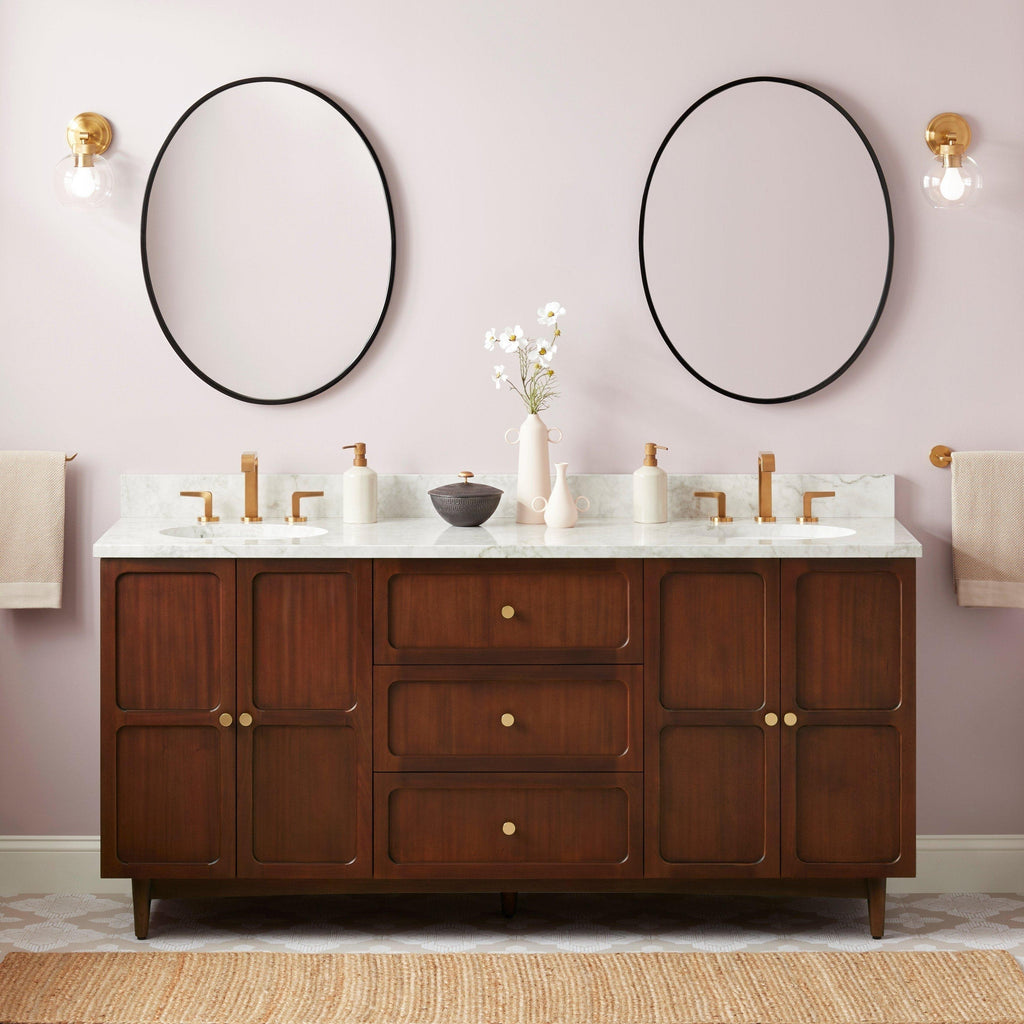 72-inch coastal solid wood double-sink vanity with Carrara marble top, two oval undermount sinks, brass faucets and knobs, round black mirrors, brass wall sconces and decorative vases against a soft pink wall.