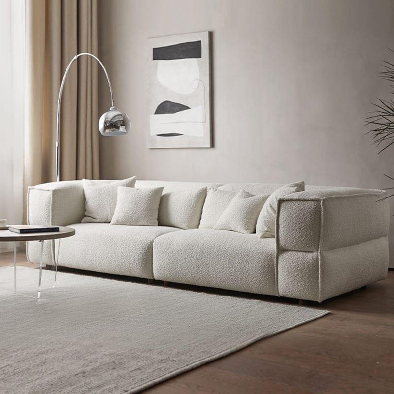 Low-profile white boucle fabric sectional sofa with plush cushions and low wooden legs in a neutral living room, styled with a chrome arc floor lamp, round coffee table, abstract wall art and textured rug.