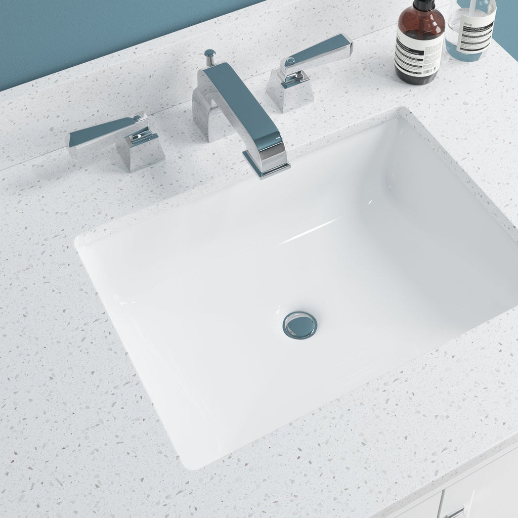 White rectangular ceramic undermount sink recessed into a speckled stone vanity top in stone color, paired with a chrome three-piece faucet and lever handles; two soap bottles rest on the back ledge against a blue wall.