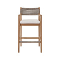 Coastal teak outdoor bar stool with a natural teak solid-wood frame, neutral woven rope back, white fabric upholstered seat cushion, armrests and a metal-reinforced footrest.