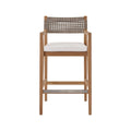 Coastal teak outdoor bar stool with a natural teak solid-wood frame, neutral woven rope back, white fabric upholstered seat cushion, armrests and a metal-reinforced footrest.