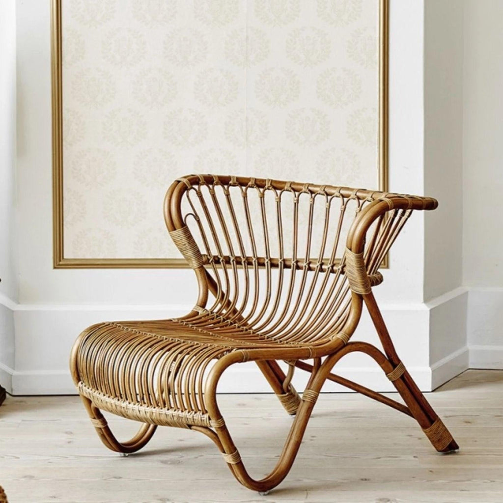 Natural rattan armless lounge chair with curved woven slat seat and back in a boho-Scandinavian style, displayed in a bright room on light wood flooring.