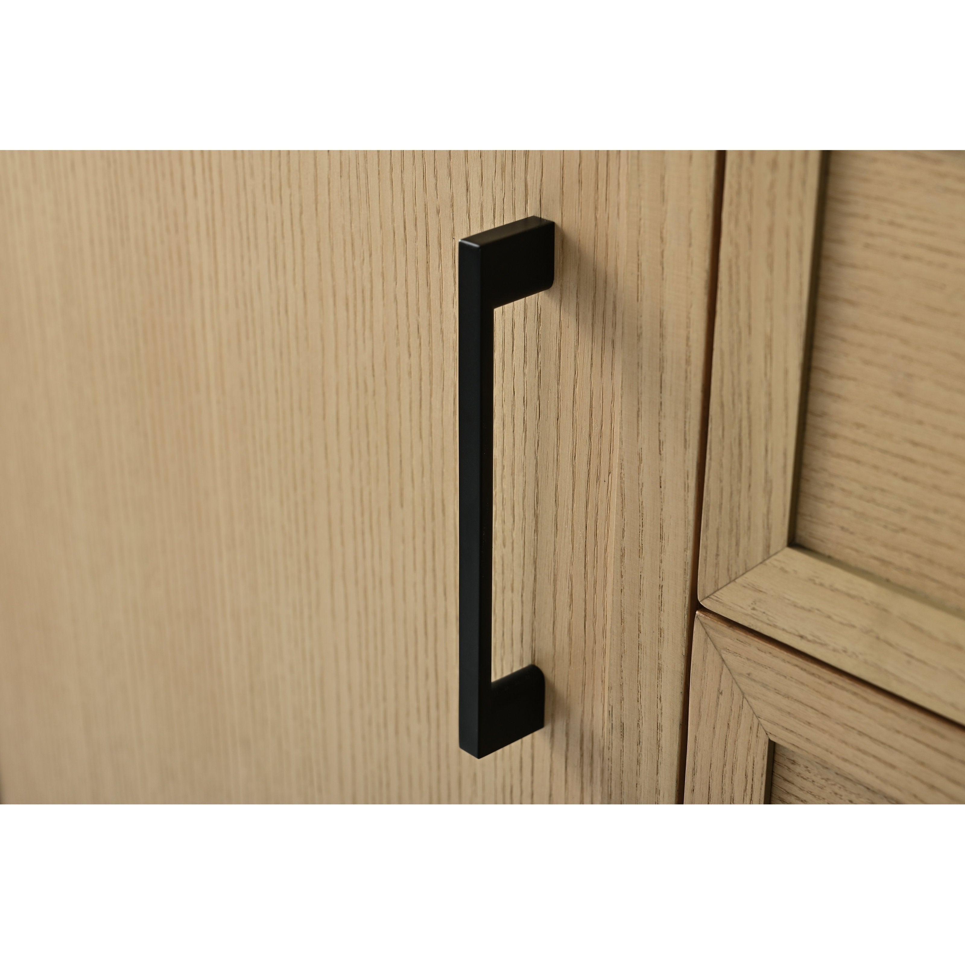 Matte black rectangular cabinet pull mounted vertically on a light stone-colored solid wood vanity door, showing wood grain texture and the adjacent recessed panel edge.