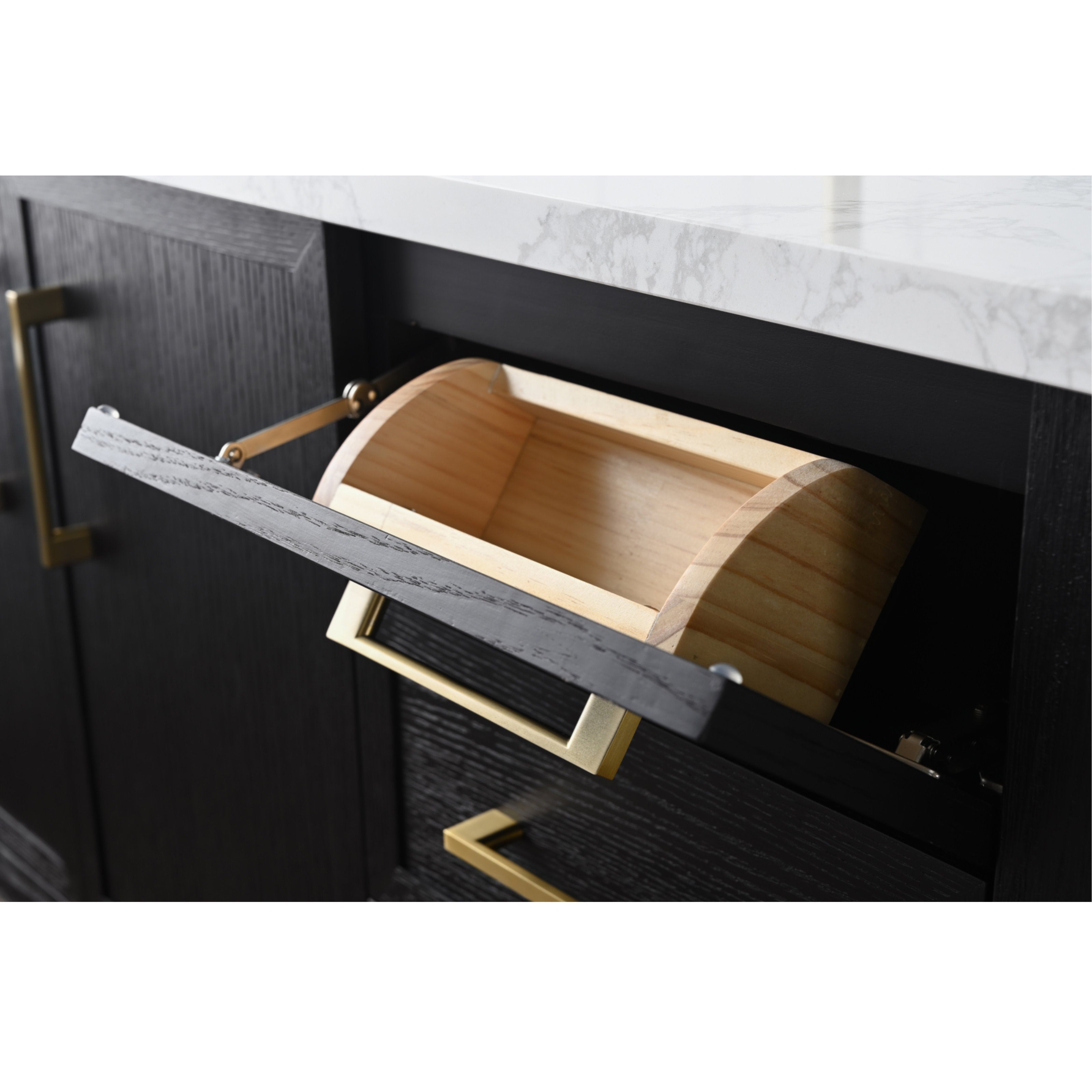 Close-up of a black solid wood bathroom vanity with white marble stone countertop, brass rectangular handles and a tilt-out front revealing a light wood curved pull-out bin for hidden storage.
