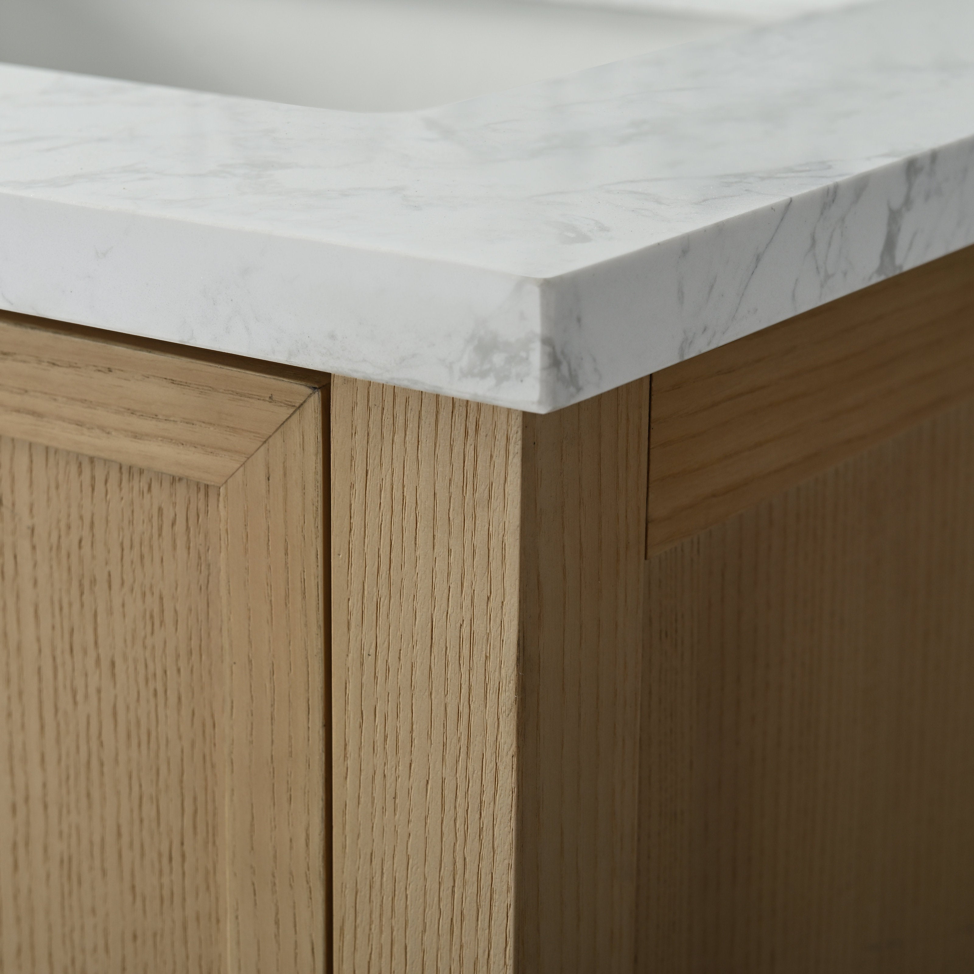 Close-up of a white marble stone top with subtle gray veining resting on a light solid wood vanity corner, showing the mitered edge and recessed panel grain detail.