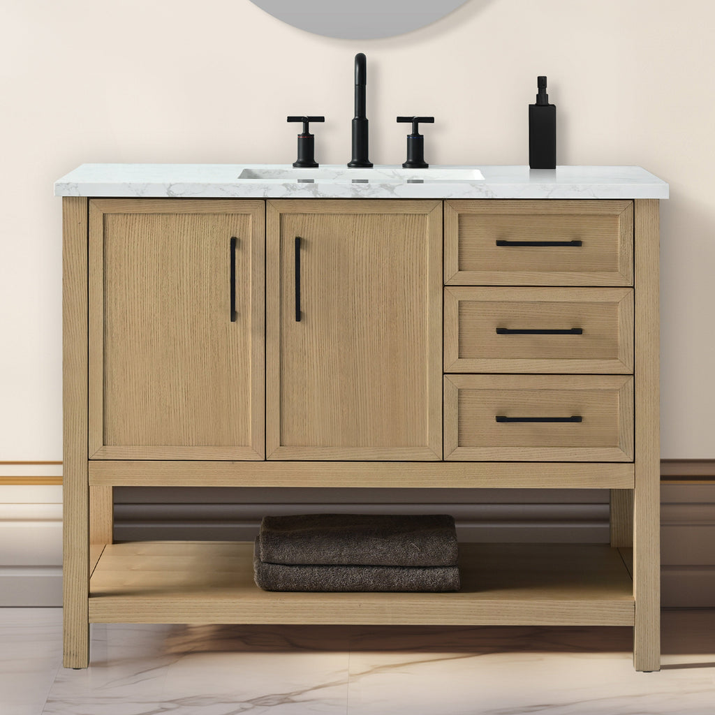 Freestanding modern single-sink bathroom vanity in light solid wood with a white marble stone top, black matte faucet and handles, undermount ceramic sink, two doors, three drawers and an open shelf with folded dark towels.
