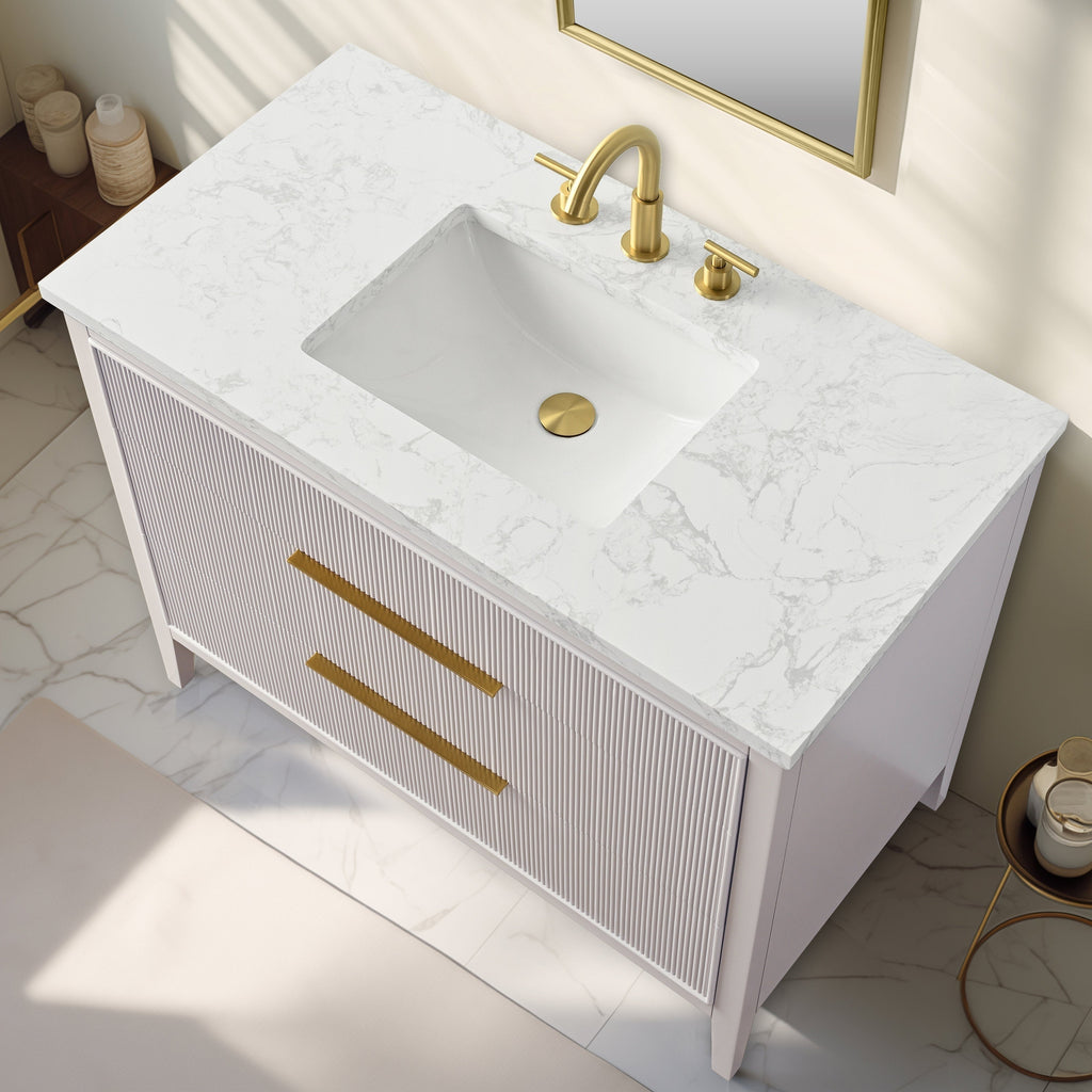 Top-down view of a modern single-sink bathroom vanity with white marbled stone top and integrated rectangular white sink, fluted light-stone solid wood cabinet, long gold drawer pulls and matching gold faucet and drain.