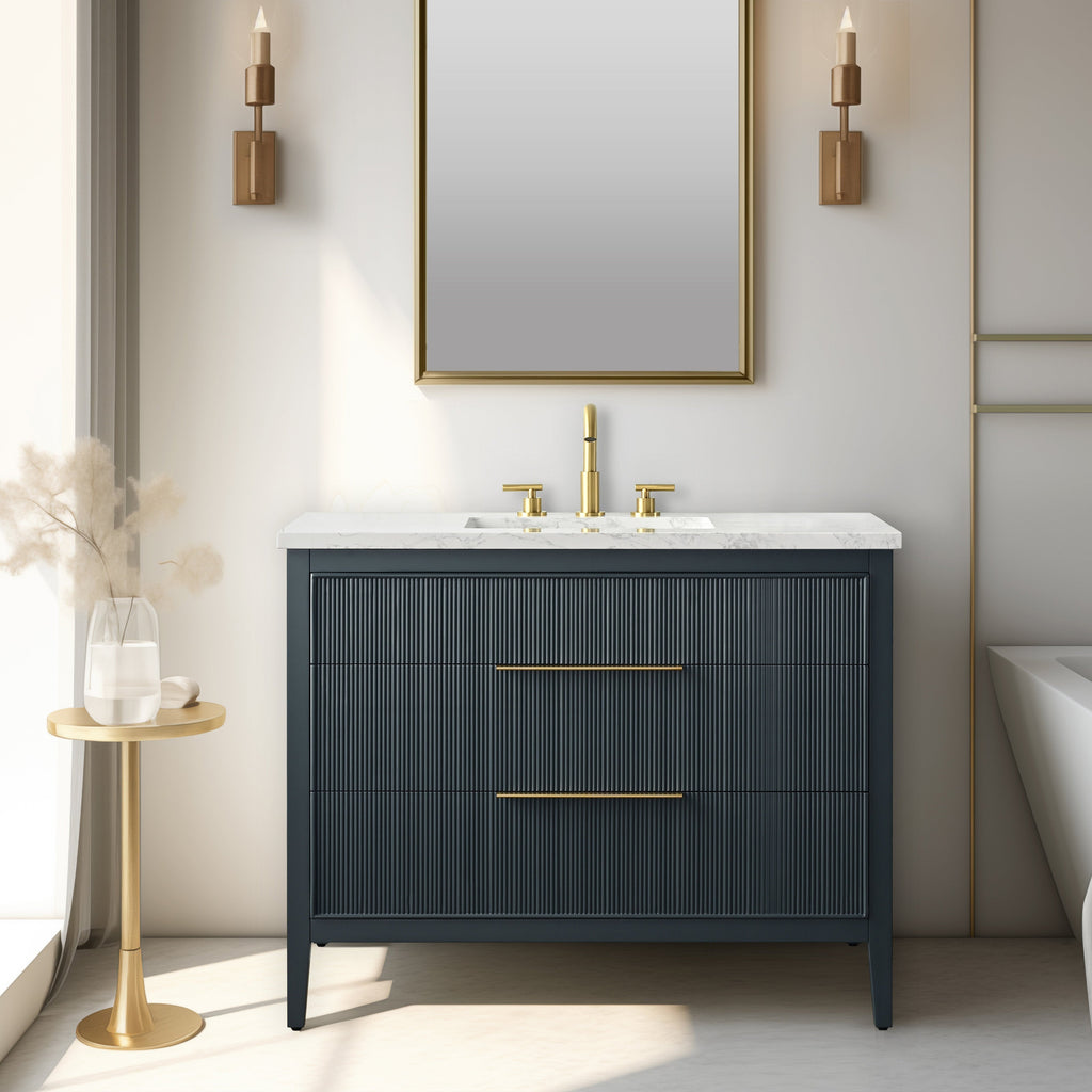 Modern solid wood single-sink bathroom vanity with a white stone top and integrated ceramic sink, dark fluted drawer fronts, brass faucet and long brass handles, freestanding tapered legs, gold-framed mirror and wall sconces.