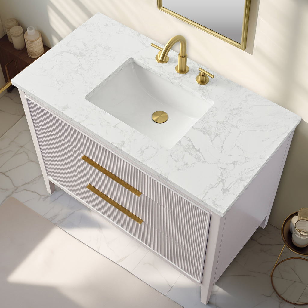 Top-down view of a modern single-sink bathroom vanity with white marbled stone top and integrated rectangular white sink, fluted light-stone solid wood cabinet, long gold drawer pulls and matching gold faucet and drain.