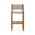 Armless teak outdoor bar stool with a natural solid-wood frame and woven beige rope seat and back, slim square legs and a front crossbar footrest in a coastal style.