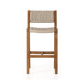 Armless teak outdoor bar stool with a natural solid-wood frame and woven beige rope seat and back, slim square legs and a front crossbar footrest in a coastal style.